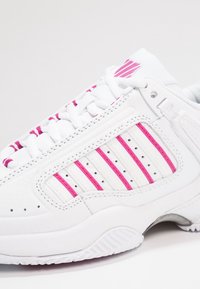 Vitaid sportskor med rosa ränder på sidan, vita snören och perforerad detaljering, visad i närbild på en vit bakgrund.