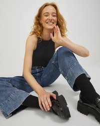 Svart ermeløs topp parret med vide blå denimbukser og svarte platå sko. Hånd hvilende på kneet, stylet hår, nøytral bakgrunn.