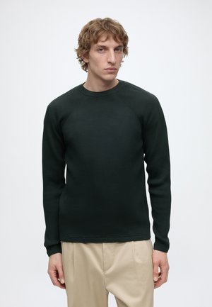Jonge man met krullend blond haar, gekleed in een aansluitende zwarte lange mouwen trui en beige broek, staand tegen een effen lichte achtergrond.