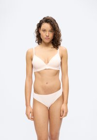 Νεαρή γυναίκα με σγουρά μαλλιά, φοράει ανοιχτό μπεζ σουτιέν και ταιριαστό εσώρουχο, κοιτώντας μπροστά με φόντο έναν απλό λευκό τοίχο.