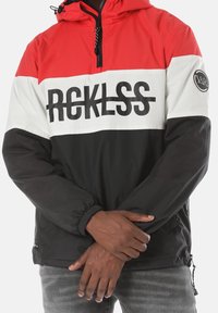 Persona che indossa una giacca colorata a blocchi nei colori rosso, bianco e nero con la scritta "RECKLESS" sul petto, con le mani incrociate davanti.