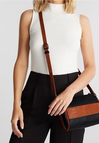 Biała prążkowana top bez rękawów z wysokim kołnierzem, zestawiony z czarnymi spodniami i małą czarną torebką z brązowym paskiem oraz detalem z logo.