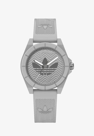 Šedé silikonové hodinky s texturovaným páskem, šestibokým pouzdrem, kulatým ciferníkem se šedým logem Adidas a bílými hodinovými a minutovými ručičkami.