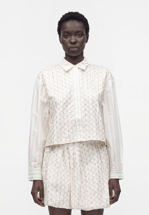 Femme aux cheveux courts et bouclés portant une chemise blanche à col et un short texturé assorti avec un motif géométrique subtil, debout devant un fond uni.