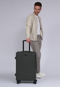 Dunkelgrüner Koffer mit Rollen im Zickzackdesign, hartschaliger Oberfläche, ausziehbarem Griff und schwarzen Rädern. Modell steht daneben.
