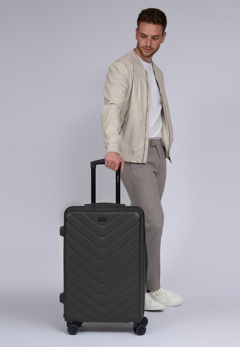 Dunkelgrüner Koffer mit Rollen im Zickzackdesign, hartschaliger Oberfläche, ausziehbarem Griff und schwarzen Rädern. Modell steht daneben.
