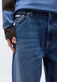 Ανοιχτό μπλε τζιν με κλασικό κόψιμο, που διαθέτει διακριτική ξεθωριασμένη υφή, λευκές ραφές και ορατή ετικέτα Calvin Klein στη μέση.