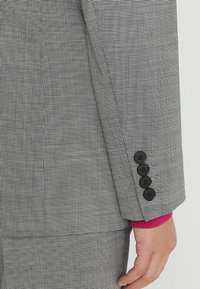 Šedá a černá kostkovaná rukáv blazera se čtyřmi černými knoflíky. Na manžetách se skrývá magentový akcent vyčnívající zpod saka.