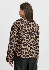 Giacca con stampa leopardata su base beige e macchie nere. Presenta una vestibilità ampia, maniche lunghe e una texture liscia. Fessure laterali sul bordo.