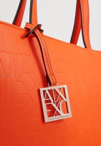 Pomarańczowa torba typu tote z silikonu z wypukłym wzorem logo, z czarnymi dodatkami i srebrnym prostokątnym zawieszką z napisem "ARMANI EXCHANGE".