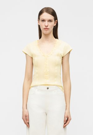 Woman with long brown hair wears a pale yellow short-sleeve top with ruffle details and white high-waisted pants against a plain background.