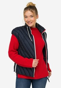 Marinblå quiltad väst med hög krage och vit dragkedja över en röd stickad tröja. Blå jeans fulländar outfiten. Vesten har inga ärmar.