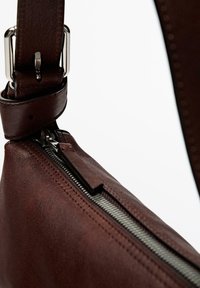 Sac en cuir brun à bandoulière avec fermeture éclair argentée, sangle ajustable et texture lisse. Présente des lignes épurées et un design minimaliste.