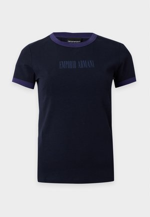 Femme aux longs cheveux foncés portant une veste en cuir marron, une chemise noire, un pantalon large foncé et des chaussures noires, debout devant un fond uni.