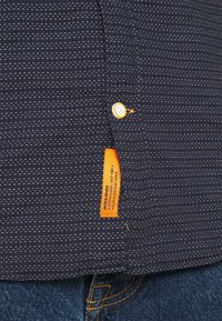 Camicia blu navale con righe bianche, dotata di un bottone giallo e un'etichetta arancione del marchio sul lato inferiore. Jeans in denim blu visibili sotto.