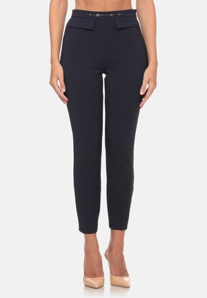 Pantaloni da donna slim fit blu navy con passanti per cintura e tasche anteriori con pattina, indossati con scarpe beige con tacco su sfondo bianco.