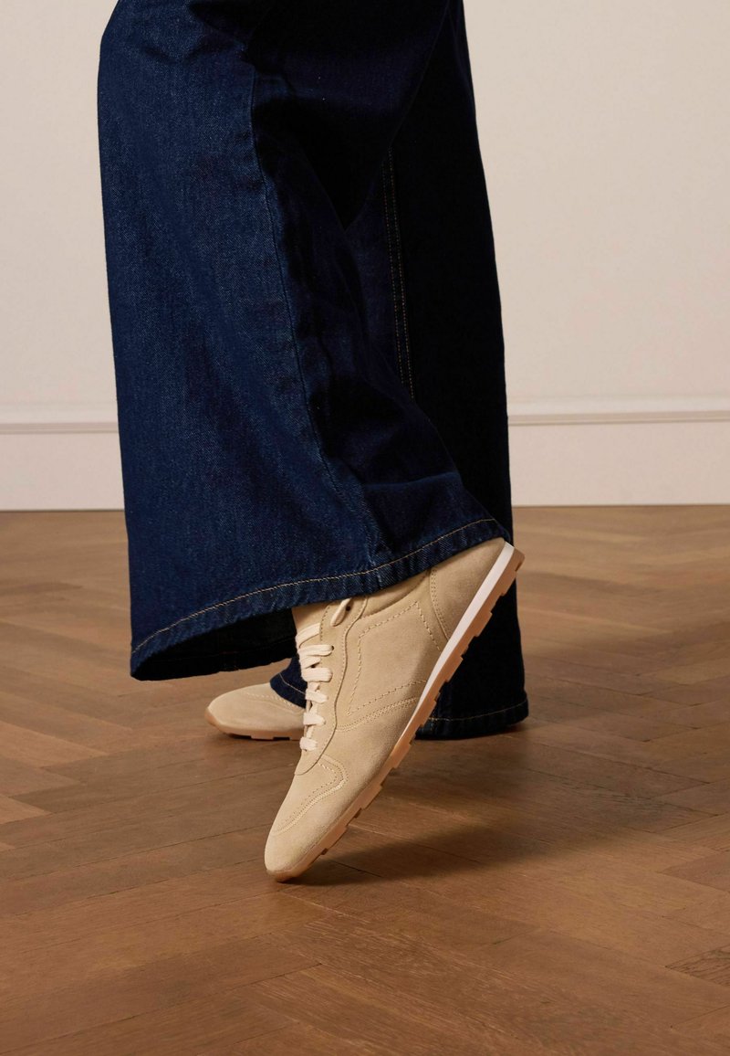 Sneaker in suede beige con lacci bianchi, punta affusolata e suola in gomma, abbinate a jeans a gamba larga blu scuro. Sfondo in legno.