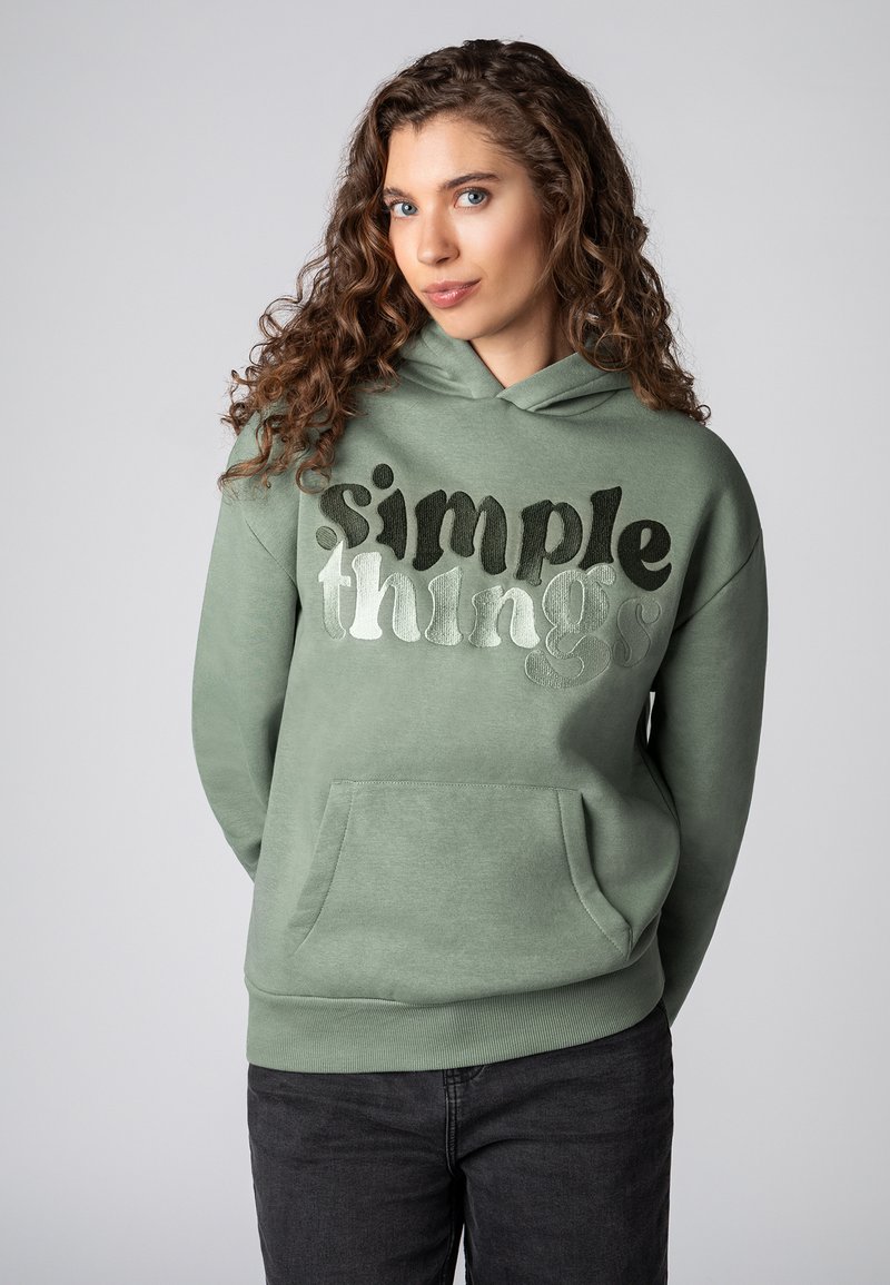 Groene hoodie met een voorzak. Bevat gestileerde tekst "simple things" in verschillende kleuren en texturen. Katoenen materiaal, relaxte pasvorm.