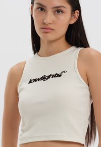 Crème-kleurige geribbelde crop top met een ronde halslijn en geborduurd "lowlights" logo in het zwart op de borst.