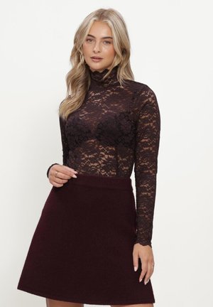 Vrouw met blond haar draagt een zwart kanten top met lange mouwen en een donkere bordeauxkleurige hoge taille rok tegen een effen achtergrond.