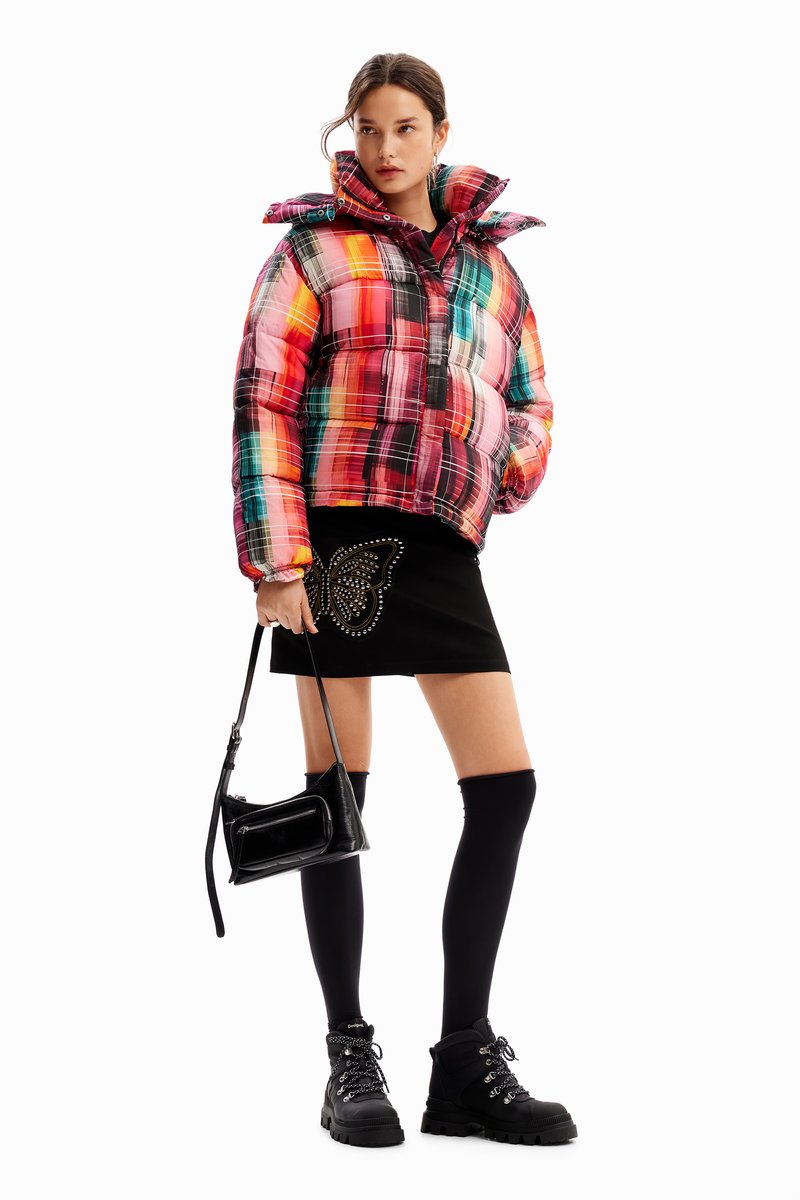 Padded, multi-colored plaid jacket with a high collar and zip closure. Black mini skirt with butterfly design, knee-high socks, and chunky boots.