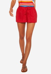 Pantalones cortos de algodón rojos con un patrón texturizado y cintura elástica con cordón. Llevados con una blusa a rayas colorida y chanclas.