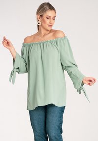 Jasnozielona bluzka na jedno ramię z falbaniastym dekoltem i długimi rękawami związanymi na mankietach, charakteryzująca się miękką teksturą i luźnym krojem.