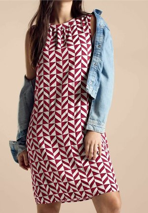 Kobieta ubrana w czerwoną i białą sukienkę bez rękawów z geometrycznym wzorem, z jeansową kurtką zarzuconą na ramiona, beżowe tło.