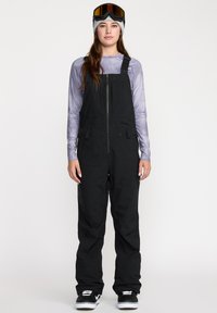 Salopettes de ski noires avec des bretelles ajustables, une fermeture éclair à l'avant et des poches. Portées avec un t-shirt à manches longues violet et des bottes de neige noires.