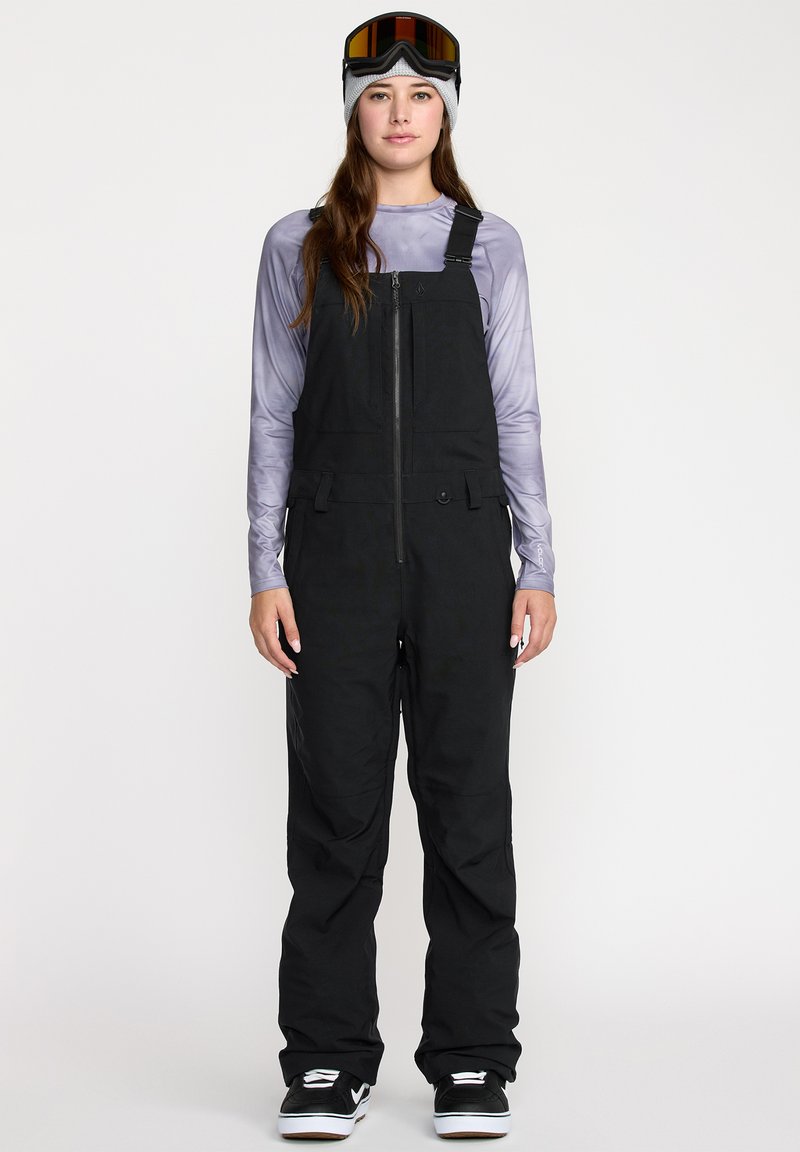 Salopettes de ski noires avec des bretelles ajustables, une fermeture éclair à l'avant et des poches. Portées avec un t-shirt à manches longues violet et des bottes de neige noires.
