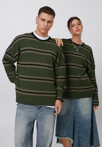 Grün gestreifte Pullover mit gerippter Textur, ausgestattet mit schwarzen und beigen Linien, kombiniert mit hellblauen baggy Jeans und einem ausgefransten Jeansrock.
