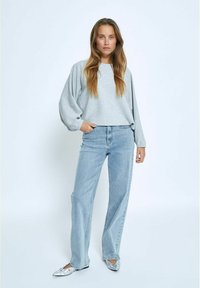 Graues Longsleeve-Oberteil mit weiten Ärmeln, kombiniert mit hellblauen, hochgeschnittenen Jeans. Das Outfit wird durch silberne, verzierte Ballerinas ergänzt.