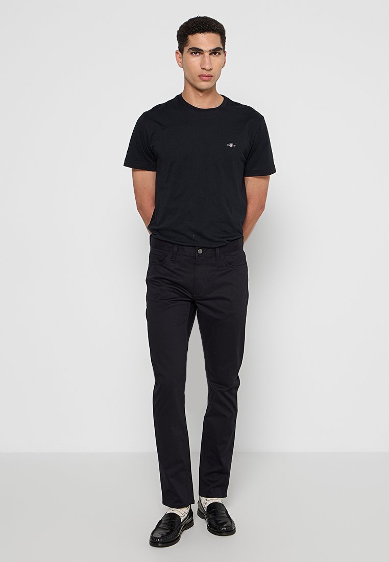 Czarna bawełniana koszulka z subtelnym logo na piersi, zestawiona z dopasowanymi czarnymi spodniami i czarnymi butami, tworząc monochromatyczny wygląd.