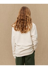 Cream-colored sweatshirt with a relaxed fit, ribbed cuffs and hem, and soft texture. Paired with dark green trousers.