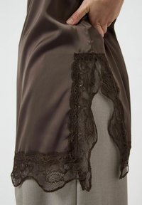 Brązowa satynowa bluzka z ozdobną koronką u dołu, o gładkiej fakturze i luźnym kroju, w połączeniu z szarymi spodniami.