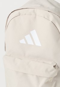 Μπεζ σακίδιο πλάτης με υφασμάτινη υφή, με λευκό λογότυπο και μπροστινή τσέπη με φερμουάρ. Ομαλές γραμμές και πρακτικός σχεδιασμός για την καθημερινή χρήση.