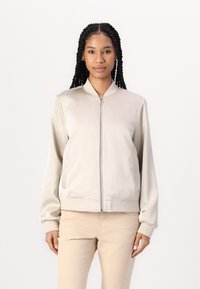 Giacca bomber beige chiaro con una superficie liscia e lucida, chiusura con zip sul davanti, polsini e bordo a costine; presenta tasche laterali e colletto alto.