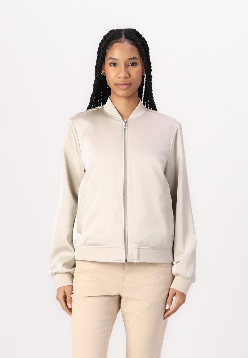 Giacca bomber beige chiaro con una superficie liscia e lucida, chiusura con zip sul davanti, polsini e bordo a costine; presenta tasche laterali e colletto alto.