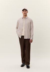 Ung mann står iført en beige stripete skjorte med knapper over en kremfarget t-skjorte, mørkebrune bukser, svarte sko og en beige caps.