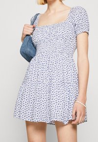 Bílé květinové šaty s modrým květinovým vzorem, hranatým výstřihem, krátkými nabíranými rukávy, staženým pasem a splývavou sukní; v kombinaci s modrou taškou.