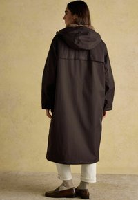 Sieviete, tērpusies garā tumši brūnā kapucē ar pūkainu apkakli, krēmkrāsas biksēs, bēšās zeķēs un brūnos mokasīnos, stāv pret vienkāršu sienu.