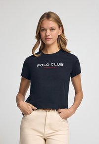 Camiseta negra de manga corta con el logo de "POLO CLUB" en texto blanco y rojo. La tela parece suave y tiene un diseño ajustado.