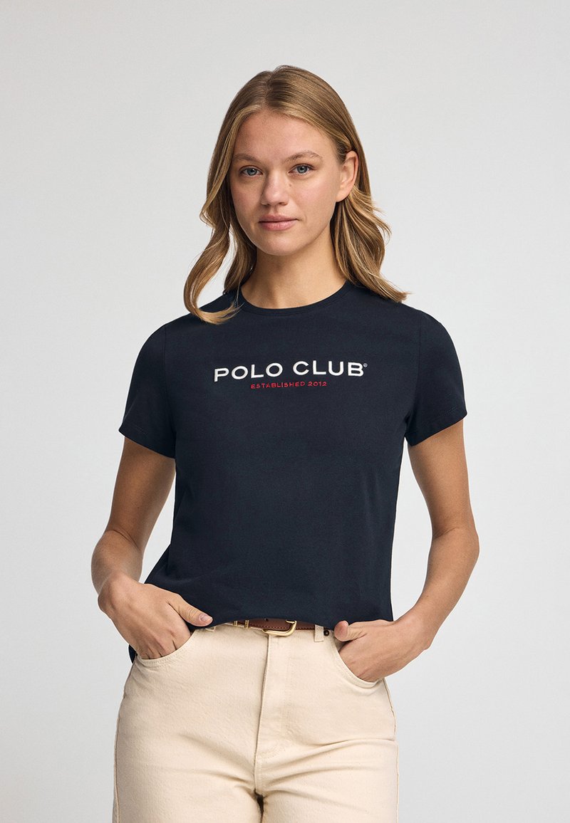 Camiseta negra de manga corta con el logo de "POLO CLUB" en texto blanco y rojo. La tela parece suave y tiene un diseño ajustado.