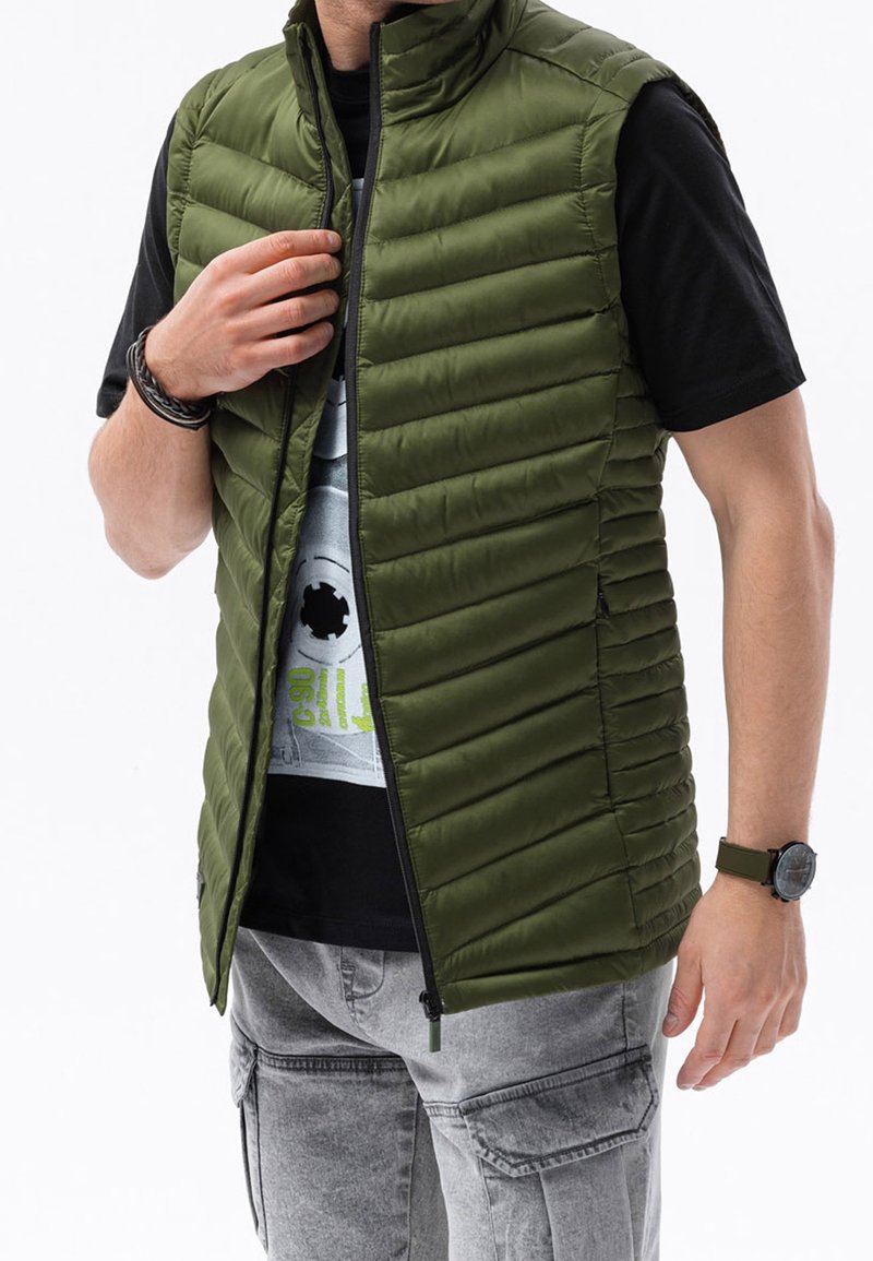 Oliwkowa marszczona kamizelka pikowana z poziomymi przeszyciami, z zamkiem błyskawicznym, wysokim kołnierzem, noszona na czarnej koszulce z nadrukiem, zestawiona z szarymi spodniami typu cargo.