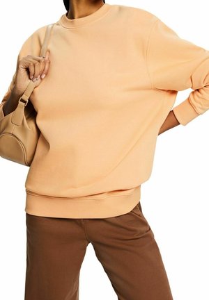 Pfirsichfarbener Sweatshirt mit Rundhalsausschnitt, langen Ärmeln und gerippten Bündchen, kombiniert mit braunen Hose und einer beigen Tasche. Weicher Stoff und lässige Passform.