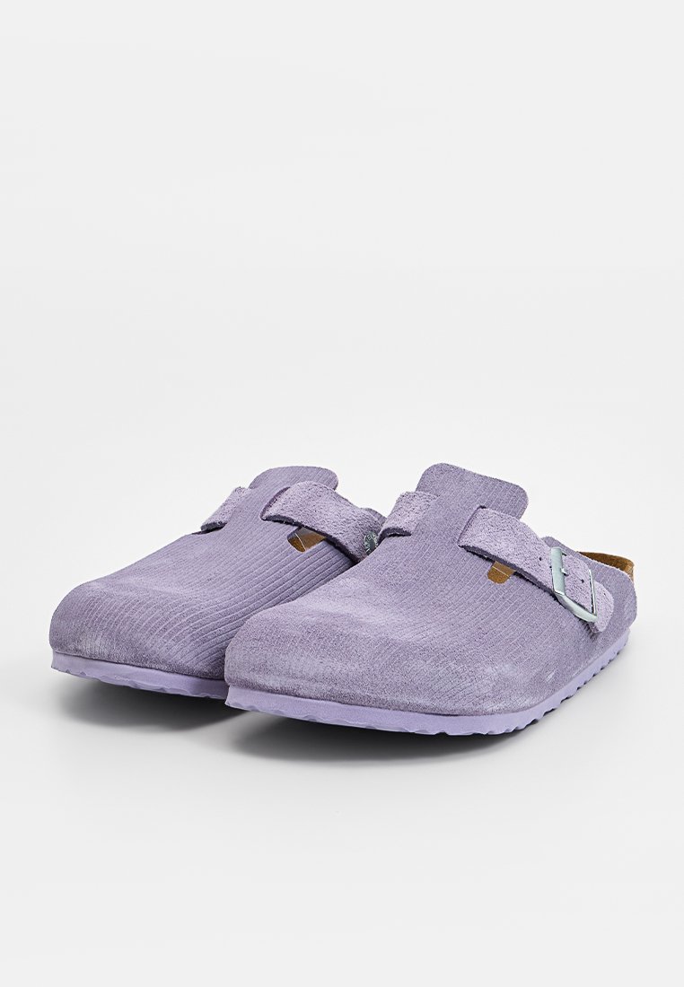 Ein Paar lavendelfarbene Wildleder-Clogs mit verstellbaren Schnallenriemen und strukturierten Sohlen, auf einem weißen Hintergrund dargestellt.