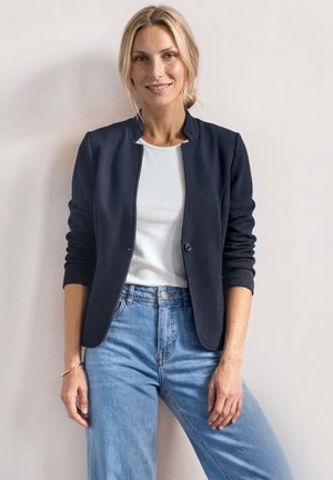 Smilende kvinde med blondt hår i en lav knold iført en marineblå blazer, hvid skjorte, lyseblå jeans og et armbånd på en lys baggrund.