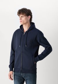Felpa con zip blu navy in misto cotone con dettaglio del logo frontale, tasche a canguro e polsini e orlo a coste, indossata su jeans in denim.