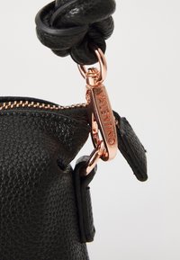 Čierna textúrovaná kožená taška so zlatým zipsom a ružovo zlatým kováním. Obsahuje uzlový kožený popruh a značkovú visačku s vyrytým logom.