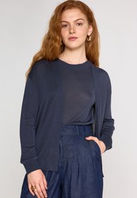 Cardigan marine avec devant ouvert, poignets côtelés et texture douce, porté par-dessus un top assorti et associé à un pantalon en denim taille haute.